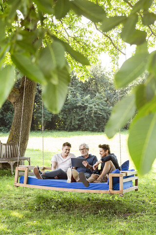 Männer einer Familie sitzen auf einem Schaukelbett im Garten, unterhalten sich, benutzen ein digitales Tablet, lizenzfreies Stockfoto