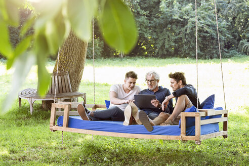 Männer einer Familie sitzen auf einem Schaukelbett im Garten, unterhalten sich, benutzen ein digitales Tablet - PESF01629
