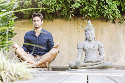 Mann sitzt im Schneidersitz neben einer Buddha-Statue in einem Zen-Garten und meditiert - PESF01630