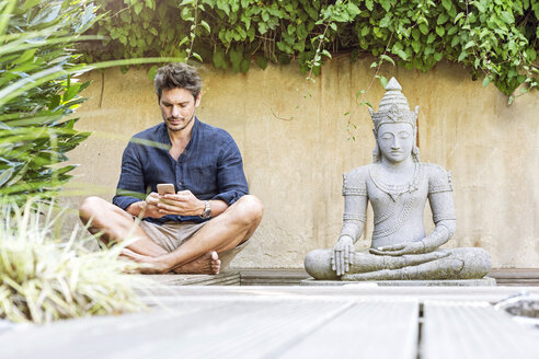 Mann sitzt im Schneidersitz neben einer Buddha-Statue in einem Zen-Garten und benutzt ein Smartphone - PESF01631