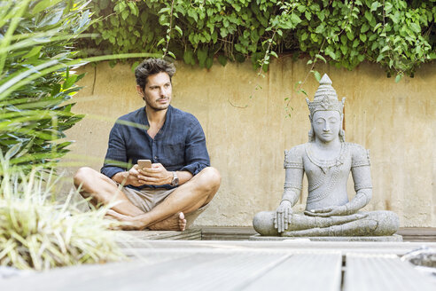 Mann sitzt im Schneidersitz neben einer Buddha-Statue in einem Zen-Garten und benutzt ein Smartphone - PESF01632