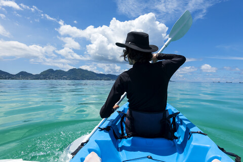Seychellen, Mahe, Rückenansicht eines schwarz gekleideten Mannes mit Hut beim Kajakfahren, lizenzfreies Stockfoto