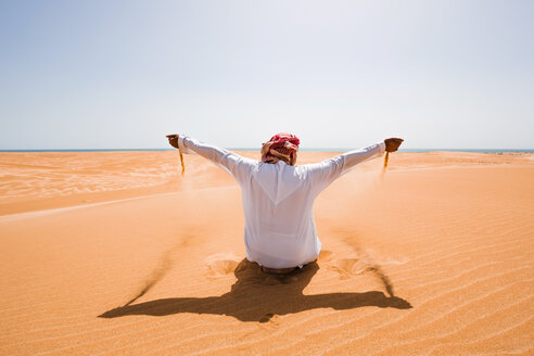 Beduine in Nationaltracht sitzt in der Wüste und hält Sand, Wahiba Sands, Oman - WVF01318