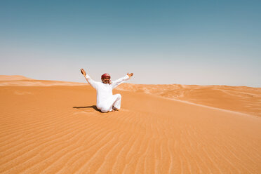 Beduine in Nationaltracht sitzt in der Wüste und hebt die Arme, Wahiba Sands, Oman - WVF01319