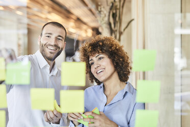 Two happy colleagues brainstorming with post-its on glass pane - FMKF05562