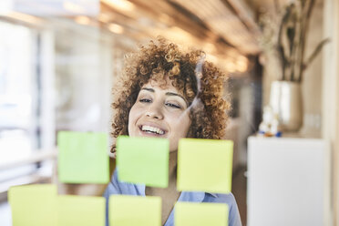 Glückliche Geschäftsfrau beim Brainstorming mit Post-its auf Glasscheibe - FMKF05567