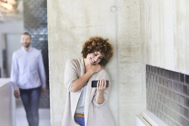 Lächelnde Frau mit Handy an Betonwand mit Mann im Hintergrund - FMKF05598