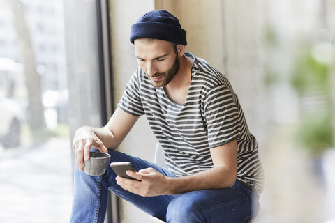 Junger Mann hält Kaffeetasse und benutzt Mobiltelefon, lizenzfreies Stockfoto