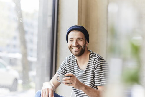 Porträt eines lächelnden jungen Mannes mit Kaffeetasse am Fenster - FMKF05609