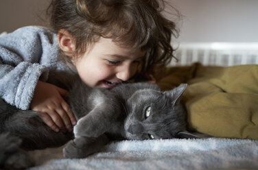 Happy toddler girl cuddling grey cat lying on bed - GEMF02920