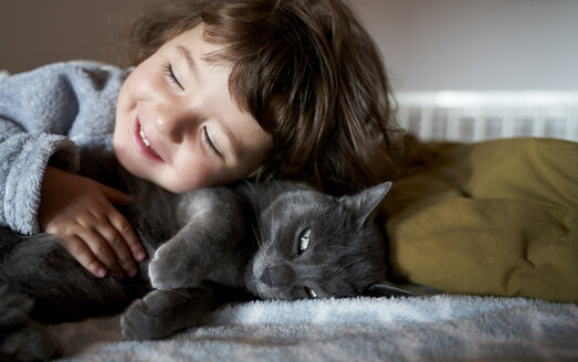 Porträt eines glücklichen Kleinkindes, das eine graue Katze auf dem Bett liegend kuschelt - GEMF02921