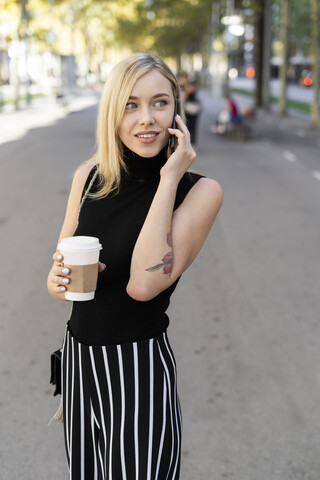 Porträt einer lächelnden blonden Frau mit Kaffee zum Mitnehmen beim Telefonieren, lizenzfreies Stockfoto
