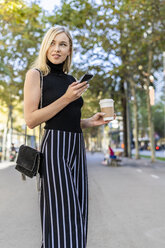 Porträt einer lächelnden blonden Frau mit Coffee to go und Mobiltelefon - GIOF06220
