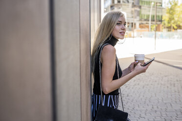 Young blond woman with coffee to go and cell phone leaning against wall looking at distance - GIOF06224