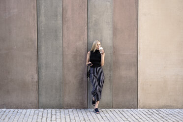 Young blond woman with cell phone leaning against wall drinking coffee to go - GIOF06225