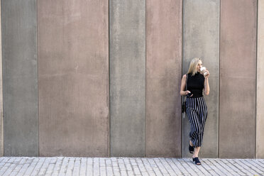 Young blond woman with cell phone leaning against wall drinking coffee to go - GIOF06226