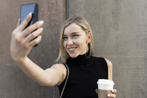 Porträt einer lächelnden blonden Frau mit Kaffee zum Mitnehmen, die ein Selfie mit ihrem Smartphone macht - GIOF06227