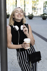Porträt einer glücklichen blonden Frau mit Coffee to go, Umhängetasche und Kopfhörern - GIOF06230