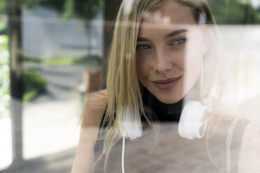 Porträt einer lächelnden blonden Frau mit Kopfhörern hinter einer Fensterscheibe - GIOF06232