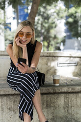 Blonde junge Frau sitzt auf einer Mauer und benutzt ein Mobiltelefon - GIOF06237