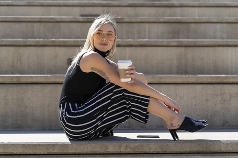 Porträt einer blonden jungen Frau mit Kaffee zum Mitnehmen, die auf einer Treppe im Freien sitzt, lizenzfreies Stockfoto