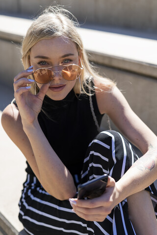 Porträt einer modischen jungen Frau, die auf einer Treppe im Freien sitzt und eine Sonnenbrille trägt, lizenzfreies Stockfoto