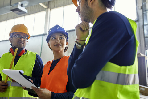 Zwei Männer und eine Frau in Warnwesten diskutieren in einer Fabrik, lizenzfreies Stockfoto