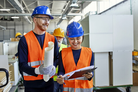 Glückliche Kollegen in Arbeitsschutzkleidung im Gespräch in der Fabrik, lizenzfreies Stockfoto