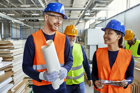 Glückliche Kollegen in Arbeitsschutzkleidung im Gespräch in der Fabrik, lizenzfreies Stockfoto