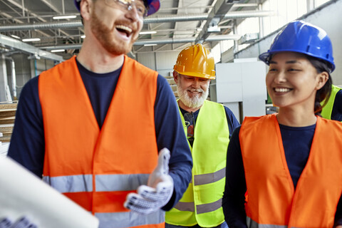 Glückliche Kollegen in Arbeitsschutzkleidung im Gespräch in der Fabrik, lizenzfreies Stockfoto