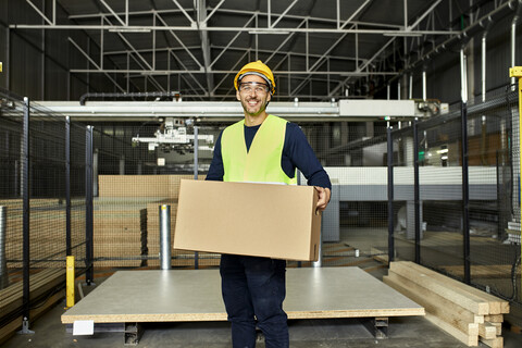 Portrait of smiling worker carrying box in factory warehouse stock photo