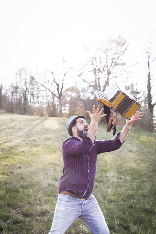 Bärtiger Mann wirft Akkordeon in die Luft auf einer Wiese, lizenzfreies Stockfoto