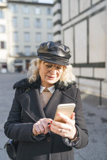 Italien, Florenz, Porträt einer reifen Frau mit schwarzem Mantel und Lederkappe, die ein Smartphone benutzt - FBAF00376
