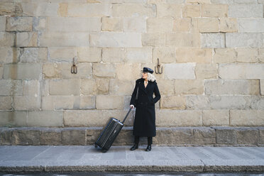 Mature businesswoman wearing black coat and leather cap standing on pavement with rolling suitcase - FBAF00381