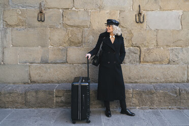 Mature businesswoman wearing black coat and leather cap standing on pavement with rolling suitcase - FBAF00382