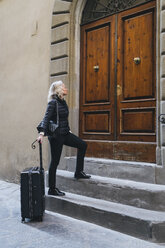 Mature woman dressed in black standing in front of entrance door with rolling suitcase - FBAF00385