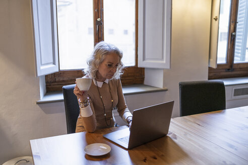 Reife Geschäftsfrau sitzt am Tisch mit einer Tasse Kaffee und benutzt einen Laptop - FBAF00388