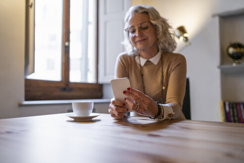 Reife Geschäftsfrau sitzt mit einer Tasse Kaffee am Tisch und benutzt ein Smartphone - FBAF00390