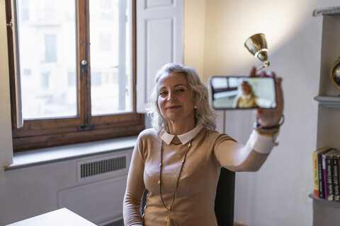 Porträt einer lächelnden reifen Geschäftsfrau, die am Tisch sitzt und ein Selfie mit ihrem Smartphone macht, lizenzfreies Stockfoto