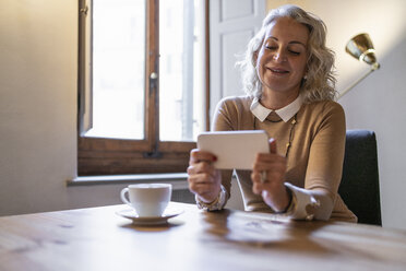 Portrait of smiling mature businesswoman sitting at table with cup of coffee looking at cell phone - FBAF00393