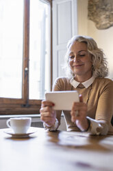 Portrait of smiling mature businesswoman sitting at table with cup of coffee looking at cell phone - FBAF00394
