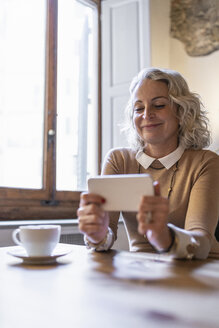 Porträt einer lächelnden reifen Geschäftsfrau, die mit einer Tasse Kaffee am Tisch sitzt und auf ihr Mobiltelefon schaut - FBAF00394