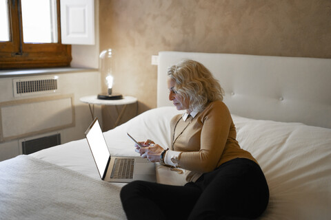 Reife Geschäftsfrau auf dem Bett liegend mit Laptop und Mobiltelefon, lizenzfreies Stockfoto