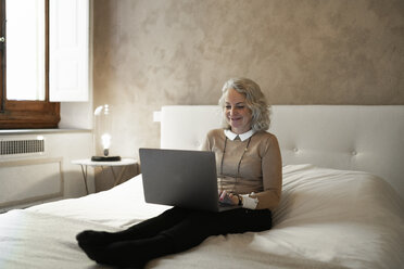Porträt einer glücklichen reifen Geschäftsfrau, die auf einem Bett sitzt und an einem Laptop arbeitet - FBAF00403