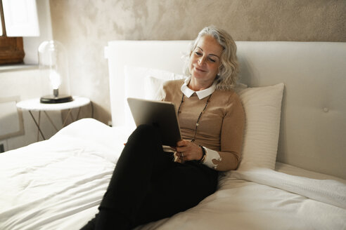 Porträt einer zufriedenen reifen Geschäftsfrau, die auf dem Bett sitzt und auf ein digitales Tablet schaut - FBAF00407