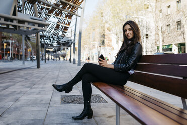 Young woman sitting on a bench, holding her smartphone - JRFF03086