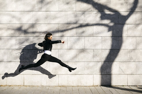 Junge Frau macht einen großen Sprung auf einer Mauer mit einem Schatten eines Baumes - JRFF03093