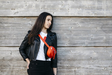 Happy young woman with a red hip bag leaning on wooden wall - JRFF03137