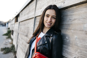 Happy young woman with a red hip bag leaning on wooden wall - JRFF03138