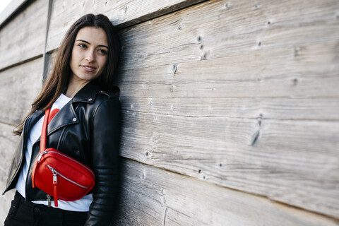 Glückliche junge Frau mit einer roten Hüfttasche, die an einer Holzwand lehnt, lizenzfreies Stockfoto